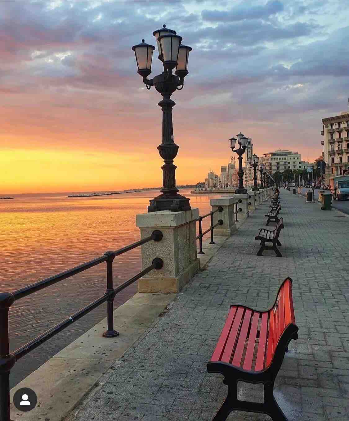 A waterfront promenade is lined with benches, offering a spot for relaxation. Elegant street lamps stand tall beside the railing. The serene water reflects the hues of a sunset sky, while charming buildings are visible in the distance.