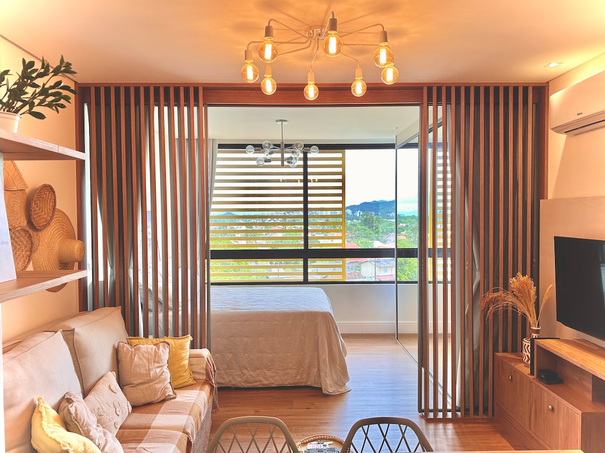 The living area features a comfortable sofa adorned with decorative cushions, complemented by wooden shelving displaying woven decor. Behind a slatted partition, a spacious bed is visible, framed by large glass windows that allow natural light and offer views of the surrounding landscape.