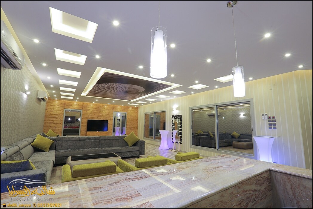 A spacious living area features multiple soft gray sofas accented with green cushions. A large marble table with a smooth surface is centered in the room. Modern ceiling lights illuminate the space, while glass doors provide a view of the outside.