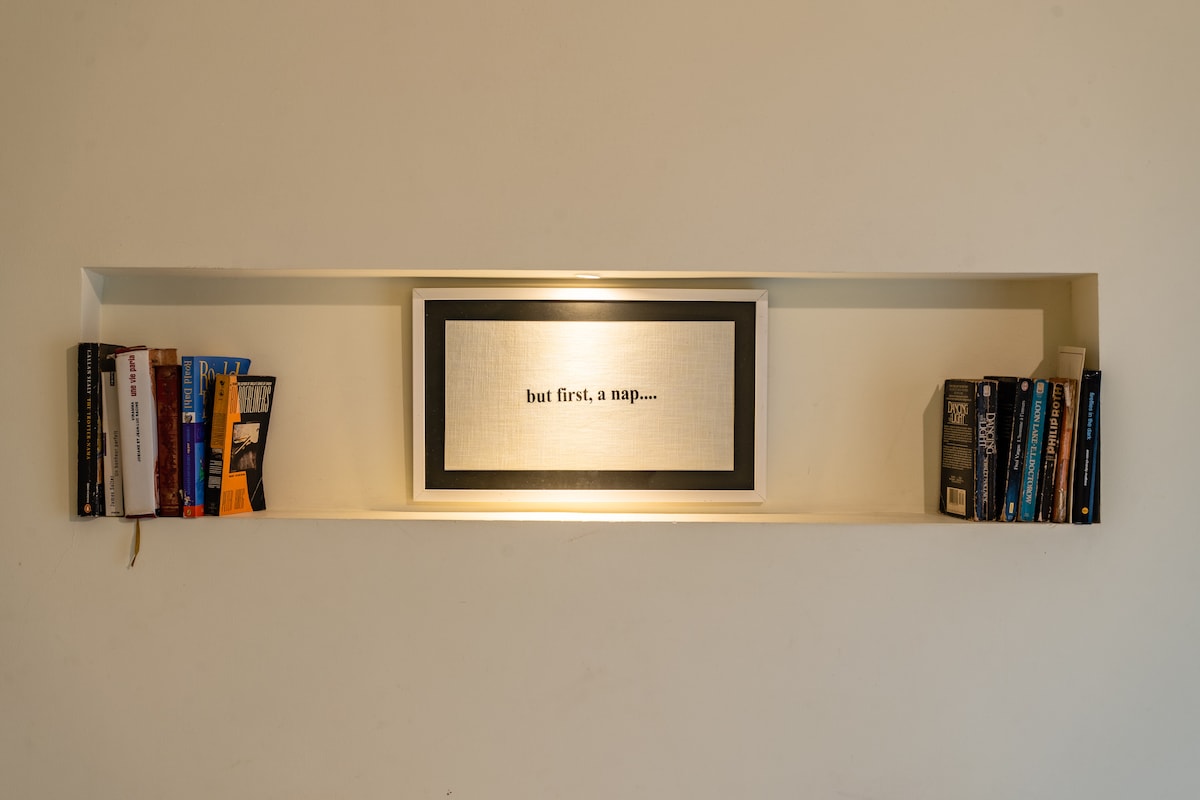 A neatly organized wall niche displays a collection of books on either side of a framed wall art piece. The artwork features the phrase 'but first, a nap...' illuminated softly, creating a cozy and inviting focal point in the space.