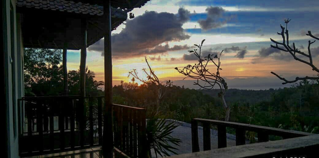 A wooden balcony offers a view of a vibrant sunset with varying shades of orange and pink over the horizon. Silhouettes of trees and distant hills are visible against the colorful sky, creating a serene backdrop.