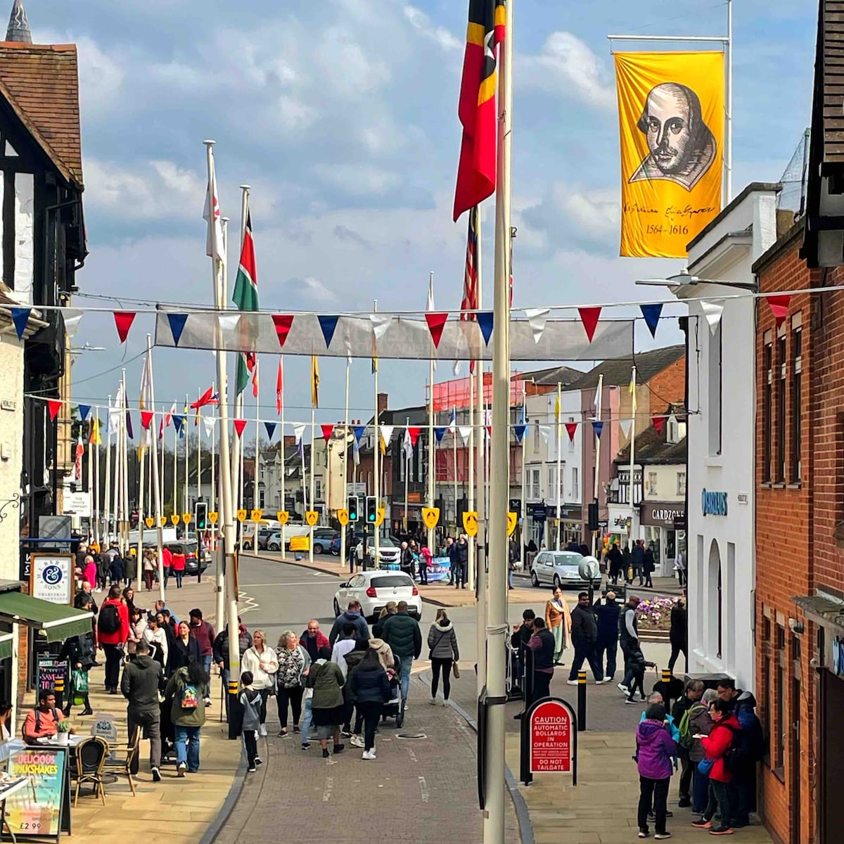 A pedestrian street bustling with activity, featuring a mix of shops and eateries. Colorful banners and flags hang above, adding vibrancy to the scene. People can be seen walking along the street, with vehicles parked nearby. The architecture reflects the historical charm of Stratford-upon-Avon.