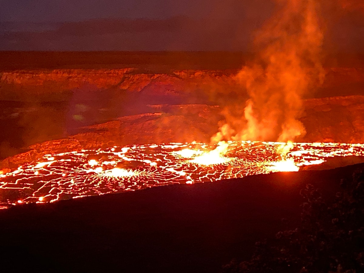 A dramatic view of an active lava flow illuminates the night sky, with glowing orange and red hues visible over the surface. Plumes of smoke rise from various points, creating a vibrant contrast against the dark landscape surrounding the volcanic crater.