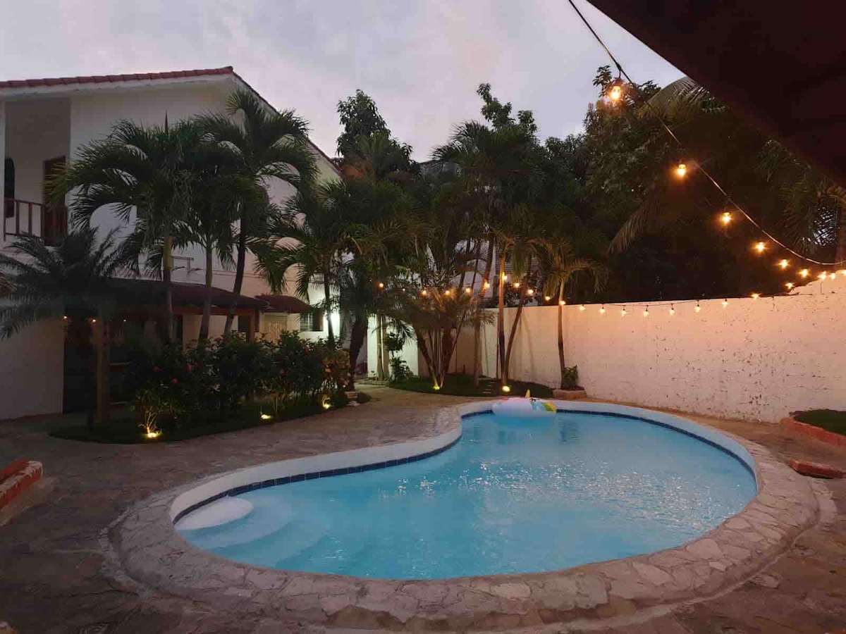 An outdoor area features a shaped swimming pool surrounded by stone tiles. Lush palm trees and plants add greenery, while string lights create a soft ambiance. A white wall provides a backdrop, contributing to a peaceful environment.