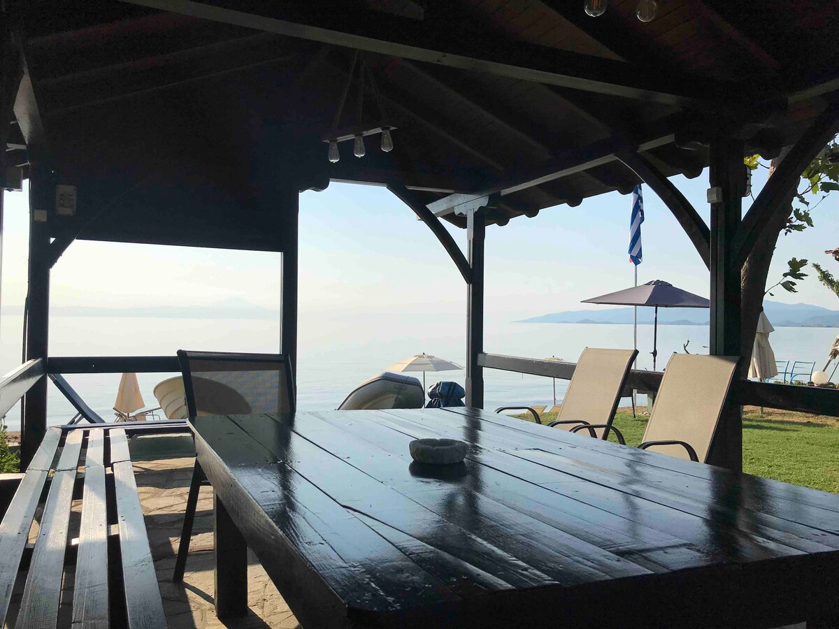 An outdoor seating area is highlighted under a wooden gazebo, featuring a large table and a pair of loungers. The serene view of the sea is visible in the background, complemented by umbrellas and sunbeds on the grassy shore.