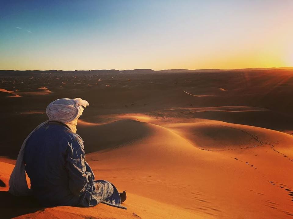A person in traditional attire sits on a sand dune, gazing at the sunset over the vast desert landscape. The shifting sands display gentle curves, while the horizon is lined with distant mountains, creating a serene and expansive view of the Sahara.