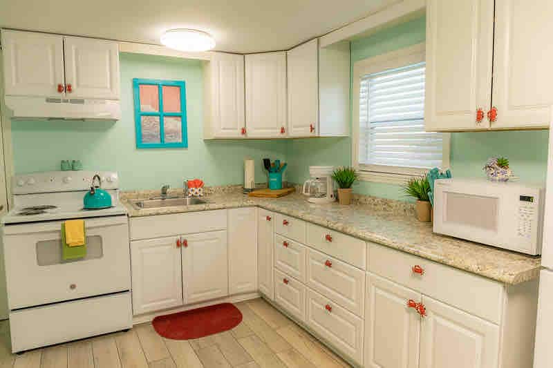 A bright kitchen area showcases light turquoise walls complemented by white cabinetry. A stove and microwave are positioned against the walls, while small potted plants add a touch of greenery. Natural light filters through a window above the sink, creating an inviting space.