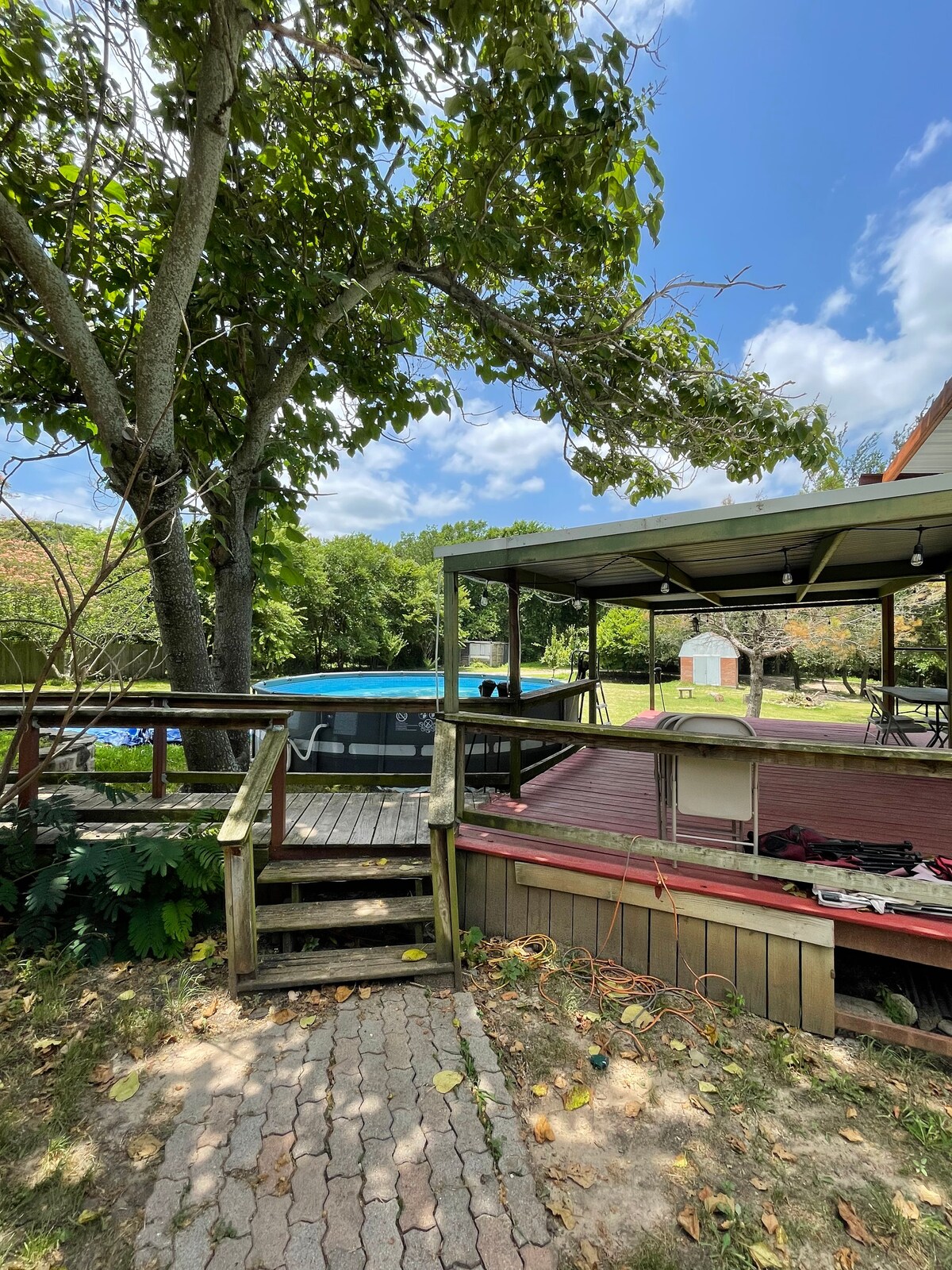 A lush outdoor area is highlighted, featuring a wooden deck next to a swimming pool. A shaded structure with seating is visible, surrounded by greenery and a paved walkway. Trees provide a natural canopy, enhancing the serene environment.