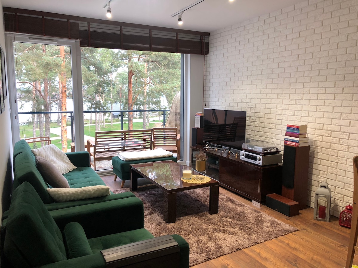 A spacious living area with green sofas is highlighted by a large glass sliding door providing views of the surrounding nature. A coffee table sits on a soft area rug, while a television and entertainment unit add functionality to the room.