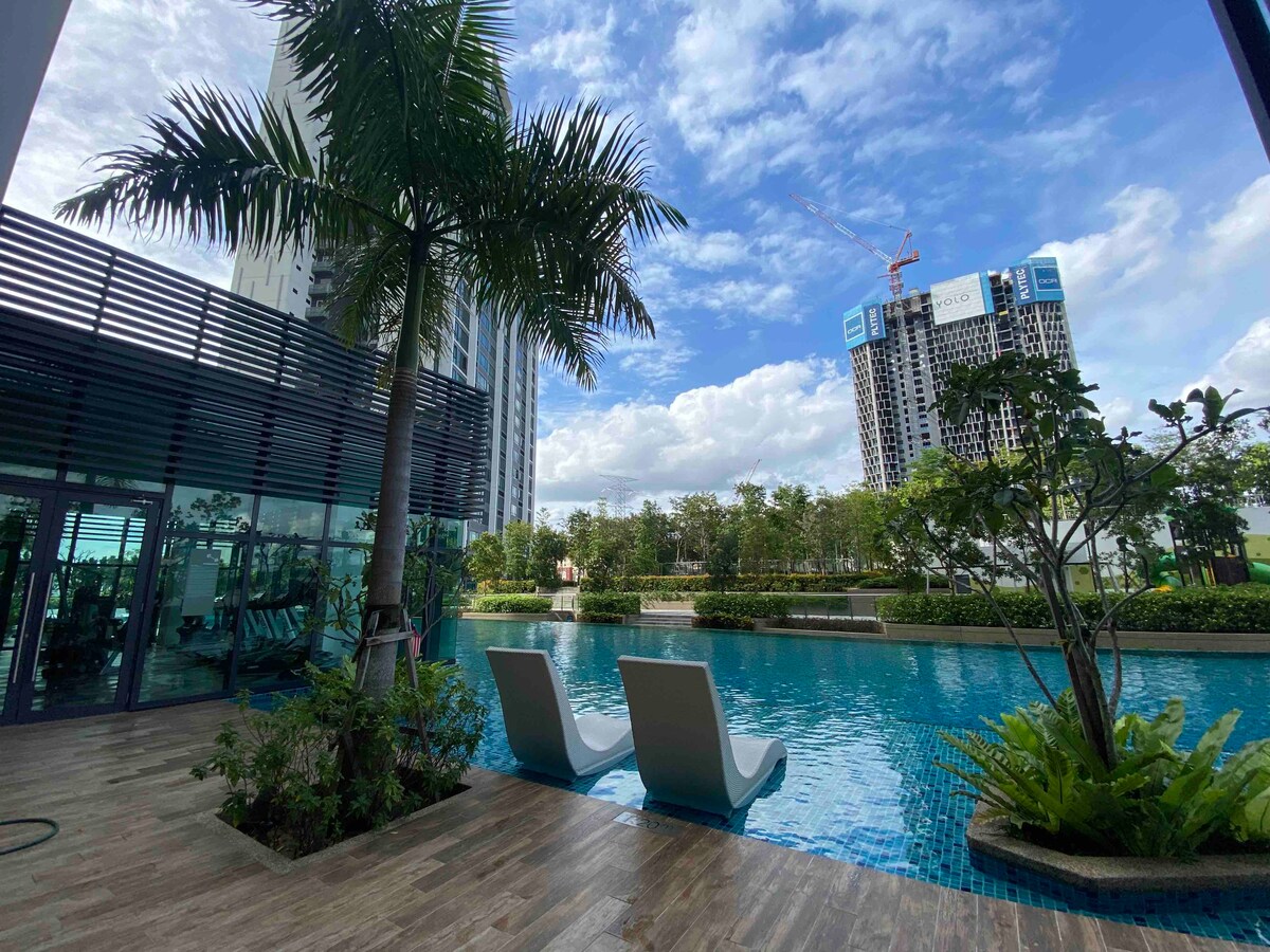 A tranquil outdoor area features a shimmering swimming pool bordered by lush greenery. Two lounge chairs are positioned by the water’s edge, offering a serene space for relaxation. In the background, modern buildings and a blue sky with scattered clouds are visible.