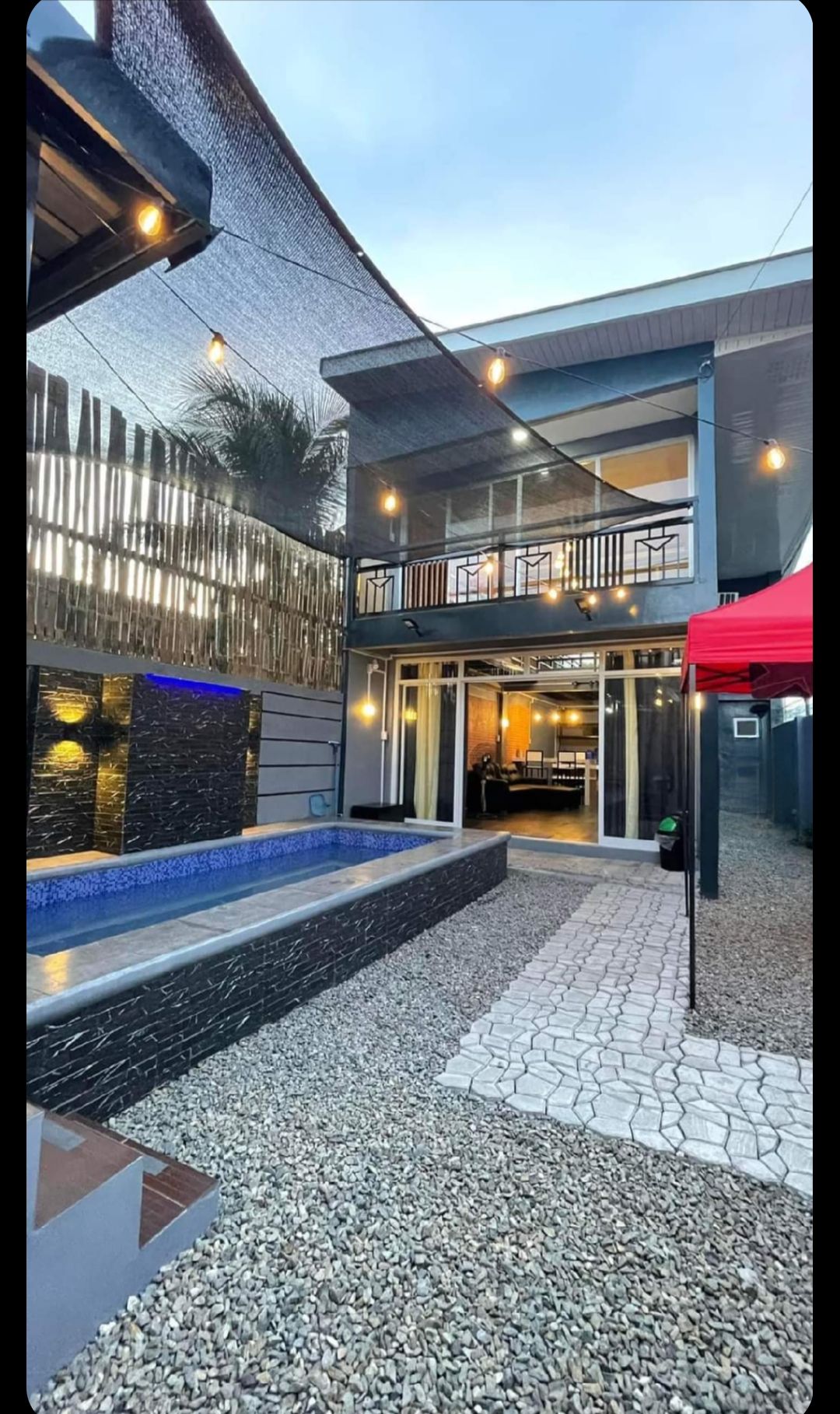 A two-story loft is depicted with expansive glass doors that open to a patio area featuring a narrow pool. Soft lighting from overhead fixtures creates a warm ambiance. The surrounding space is enhanced with pebble-stone pathways and shaded areas, providing a serene outdoor experience.