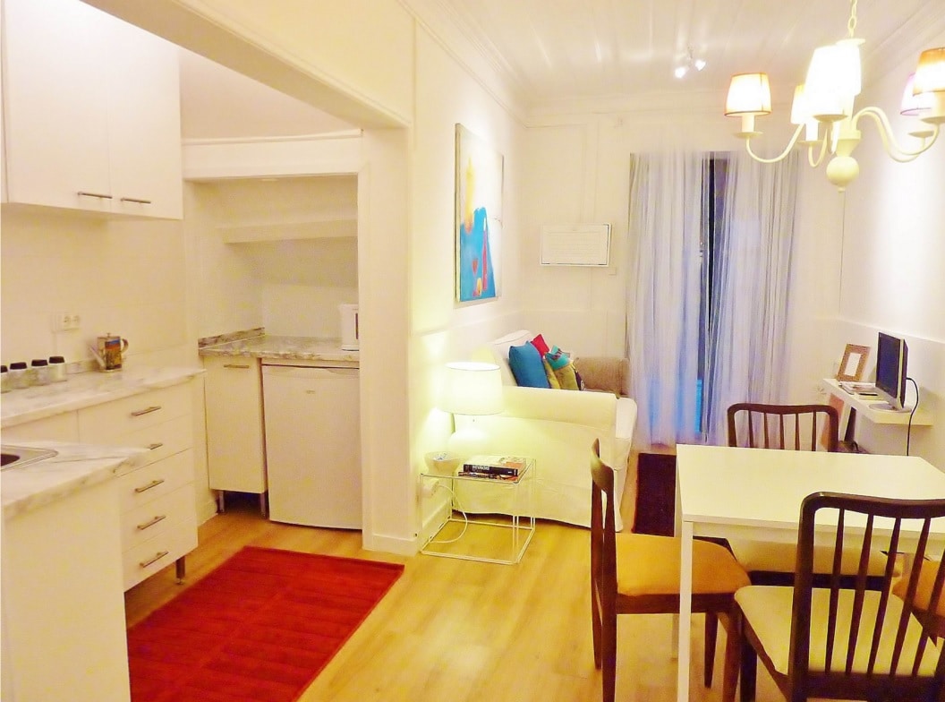The cozy kitchenette is equipped with modern appliances, including a refrigerator and stove, while a small dining area with a white table and chairs is visible. A comfortable sofa is placed in the living area, with soft lighting provided by a stylish chandelier overhead.