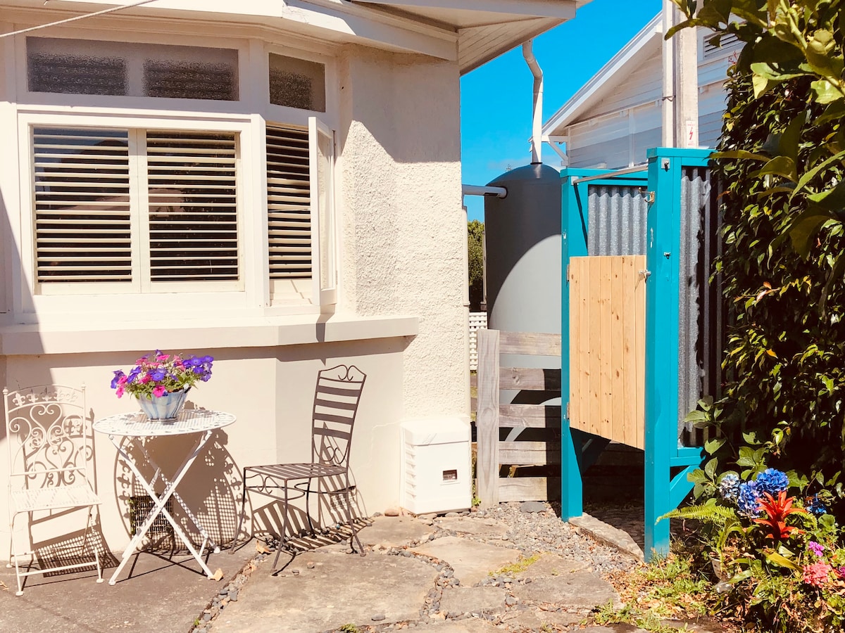 A small outdoor courtyard is shown, featuring a round table with two chairs beside a colorful flower pot. Textured walls and greenery surround the space, providing a tranquil atmosphere. An outdoor shower is visible, alongside a pathway leading to the property entrance.