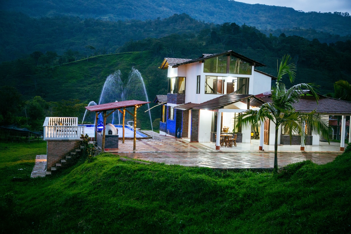 A modern two-story house is positioned against a backdrop of lush mountains. Water features are gently illuminated, adding a serene quality. An outdoor dining area is visible on the spacious patio, while expansive windows invite natural light into the interior.
