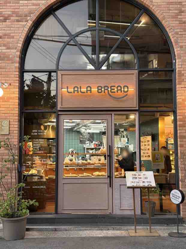A bakery facade features a large arched window with a wooden door, displaying fresh baked goods inside. Potted plants are placed near the entrance, and a menu sign stands on the sidewalk. The brick exterior adds to the warm and inviting atmosphere.