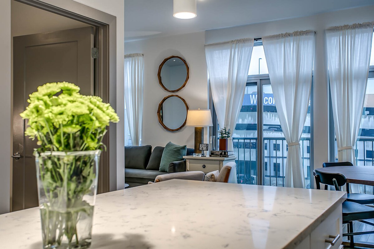 A bright living area features large windows adorned with sheer curtains that allow natural light to fill the space. A gray couch and a wooden side table are visible, complemented by decorative mirrors on the wall. A bouquet of green flowers is placed in a glass vase on a marble countertop.