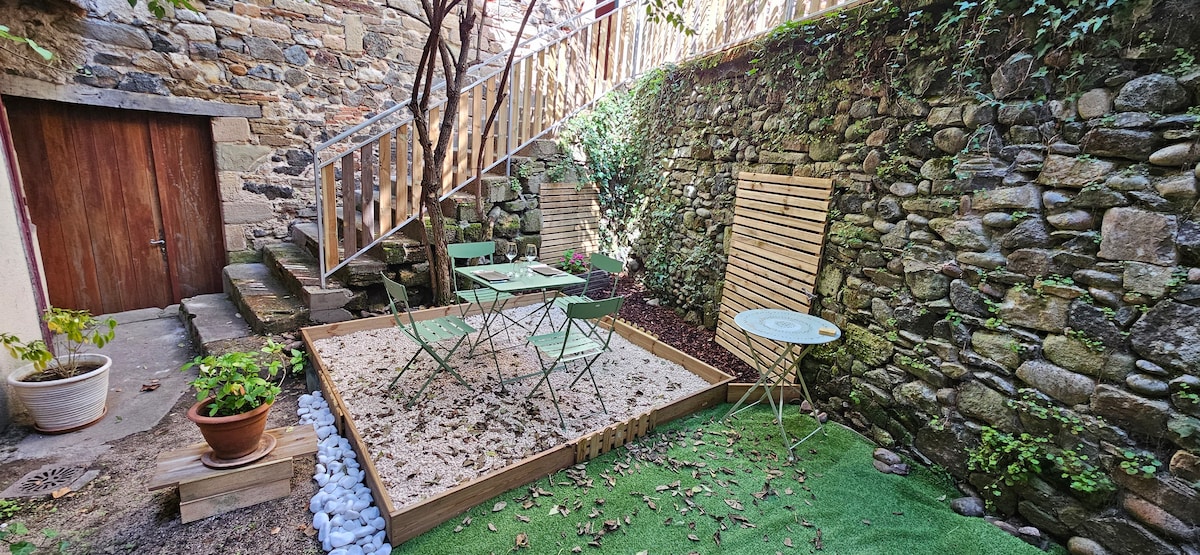 A small outdoor space features a sandy area with four green folding chairs and a round table. Natural stone walls line the area, complemented by a wooden structure in the background. Potted plants and decorative stones add a touch of greenery.