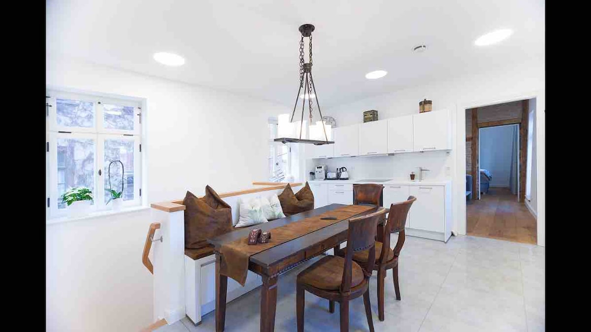 A clean and well-lit kitchen area features a modern dining table made of wood, accompanied by several wooden chairs. Illuminated by a stylish pendant light, the space showcases a wall-mounted kitchen with ample storage and appliances. Large windows provide natural light, enhancing the inviting atmosphere.