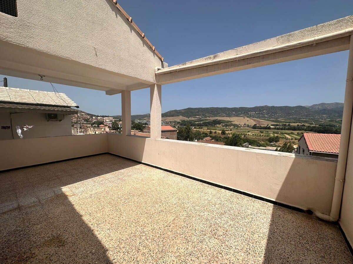 An expansive terrace offers sweeping views of the surrounding countryside. Sunlight casts subtle shadows on the textured floor, highlighting the spacious outdoor area. The backdrop includes rolling hills and distant mountains under a clear blue sky.