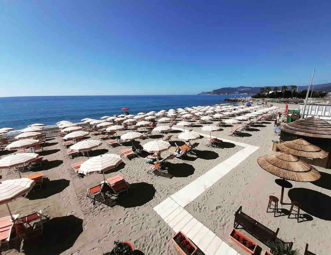A wide sandy beach is lined with numerous umbrellas and sun loungers, offering ample relaxation space. The serene blue sea stretches into the distance under a clear sky, while a few distant hills are visible along the shore.