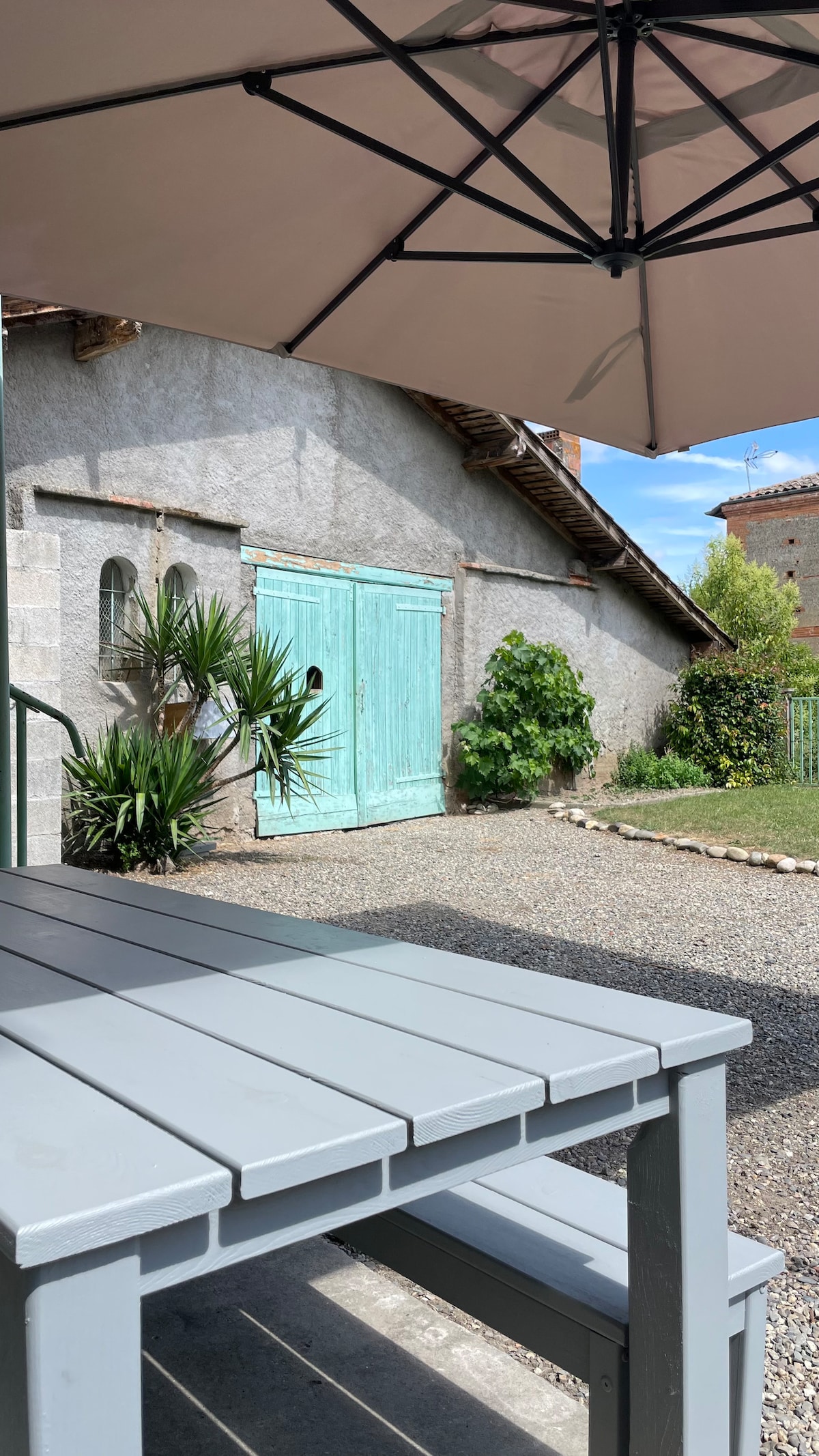 A gravel patio offers a shaded dining area beneath a large umbrella. In the background, a light blue door adds a pop of color to the stone wall. Lush greenery and a small garden are visible, contributing to a serene outdoor environment.