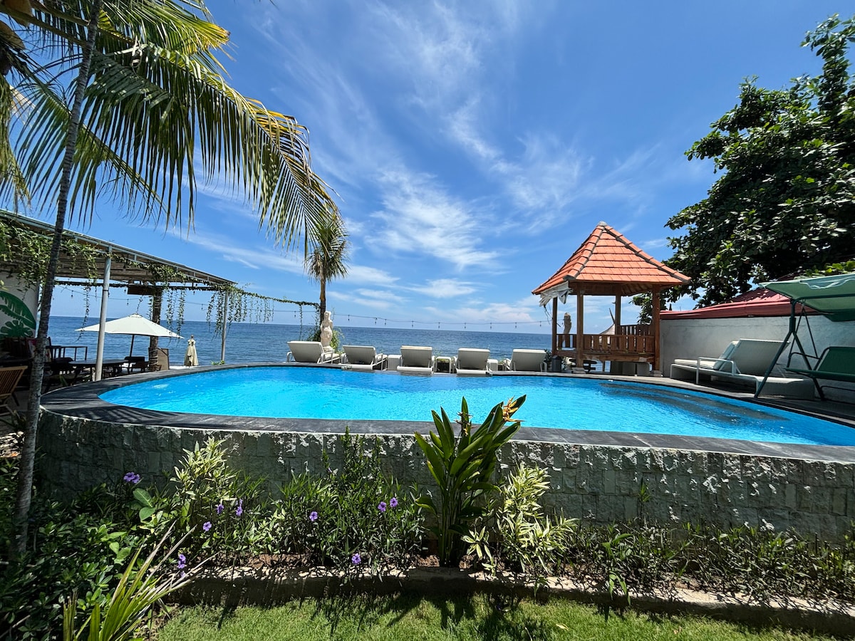 A tranquil swimming pool features clear blue water, surrounded by greenery and lounge chairs. In the background, a traditional gazebo is seen alongside the ocean. Bright skies and gentle waves complement the serene environment.