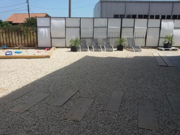 A gravel area is shown with wooden walkways leading through. Sun loungers are positioned on one side, accompanied by potted plants. A fence is visible in the background, along with a brightly colored play area and a structure featuring large windows.