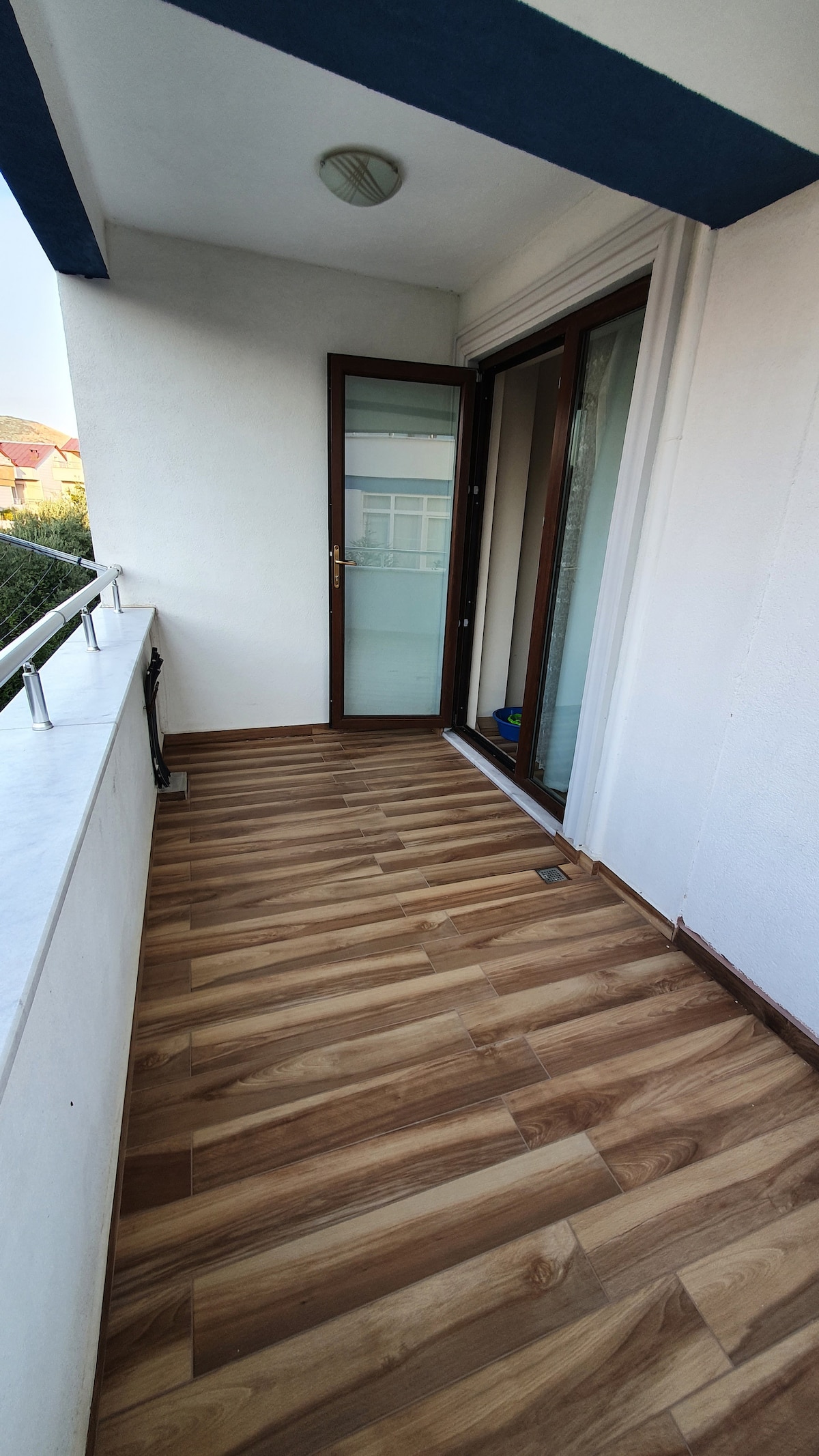 A small balcony is shown, featuring wooden flooring and a white wall. Sliding glass doors provide access to an interior space, with a light fixture mounted on the ceiling. A railing runs along the perimeter, and a glimpse of greenery can be seen beyond the balcony.