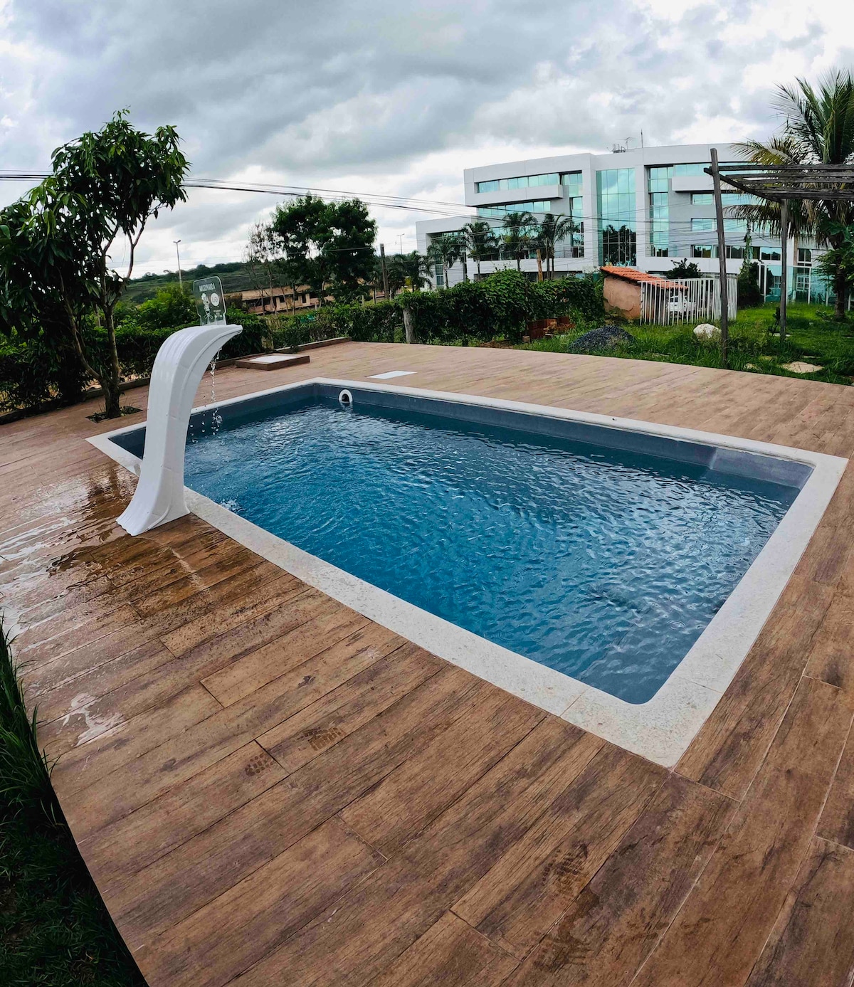 A rectangular swimming pool is set on a wooden deck surrounded by lush greenery. A water feature gracefully cascades into the pool, while modern buildings can be seen in the background. The scene is shaded by overcast skies.
