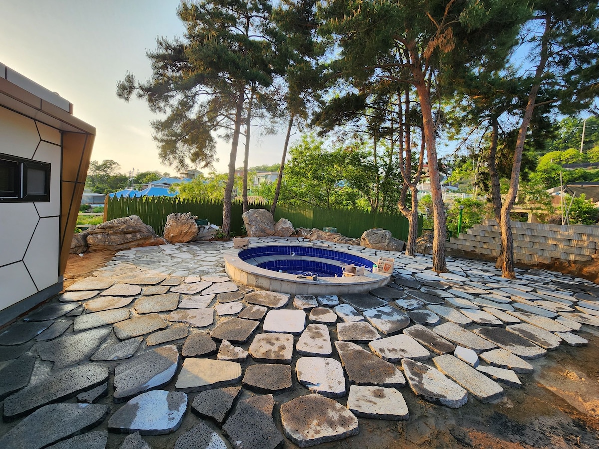 A circular hot tub is situated on a stone patio, surrounded by tall pine trees. The area features a mixture of large gray and white stones. Green foliage and a wooden fence are visible in the background, enhancing the natural setting.