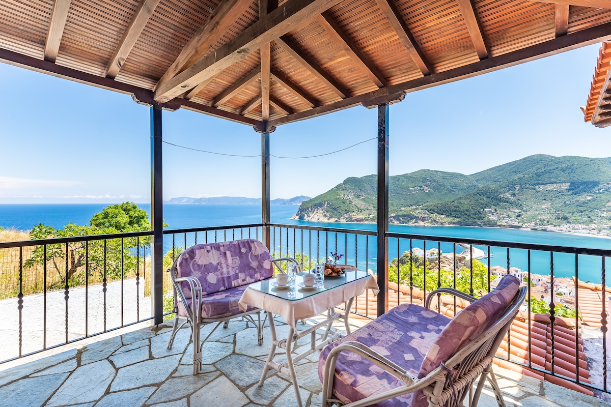 A shaded terrace features two cushioned chairs and a small table, offering views of the Aegean Sea and surrounding hills. The wooden ceiling adds warmth, while the stone floor enhances the outdoor setting. A railing offers a safety barrier while allowing for unobstructed vistas.