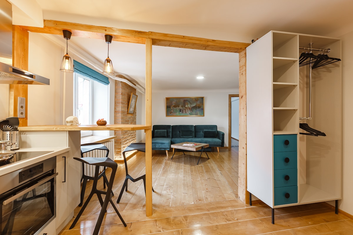 A combined living and dining area features a stylish seating arrangement with a teal sofa and a minimalist coffee table at the center. Natural light fills the space through a window, and a well-equipped kitchen with modern appliances is visible in the foreground, complemented by wooden accents.