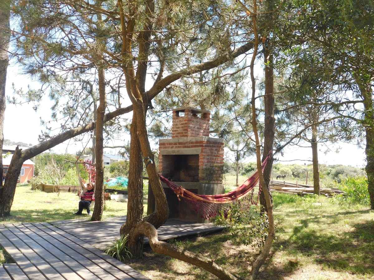 A cozy outdoor space is framed by tall pine trees, featuring a brick barbecue grill beside a hammock. Wooden decking provides a relaxing area under the natural canopy, with greenery visible in the background, creating a serene setting for outdoor gatherings.