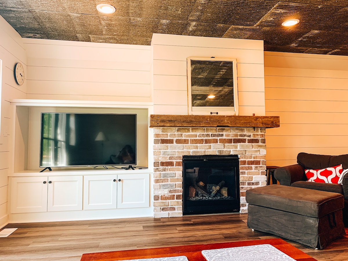 A comfortable living area features a wooden mantle above a brick fireplace, with a sleek television housed in a white media unit. Soft lighting highlights the space, and a dark sofa with decorative cushions invites relaxation, creating a cozy atmosphere.