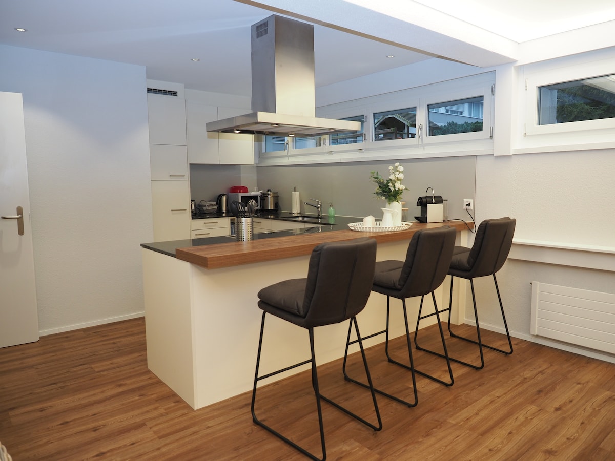 A modern kitchen is presented with light cabinetry and a sleek countertop. Four high bar stools are arranged around the island, providing casual seating. Large windows allow natural light to illuminate the space, enhancing the functionality of the appliances visible in the background.