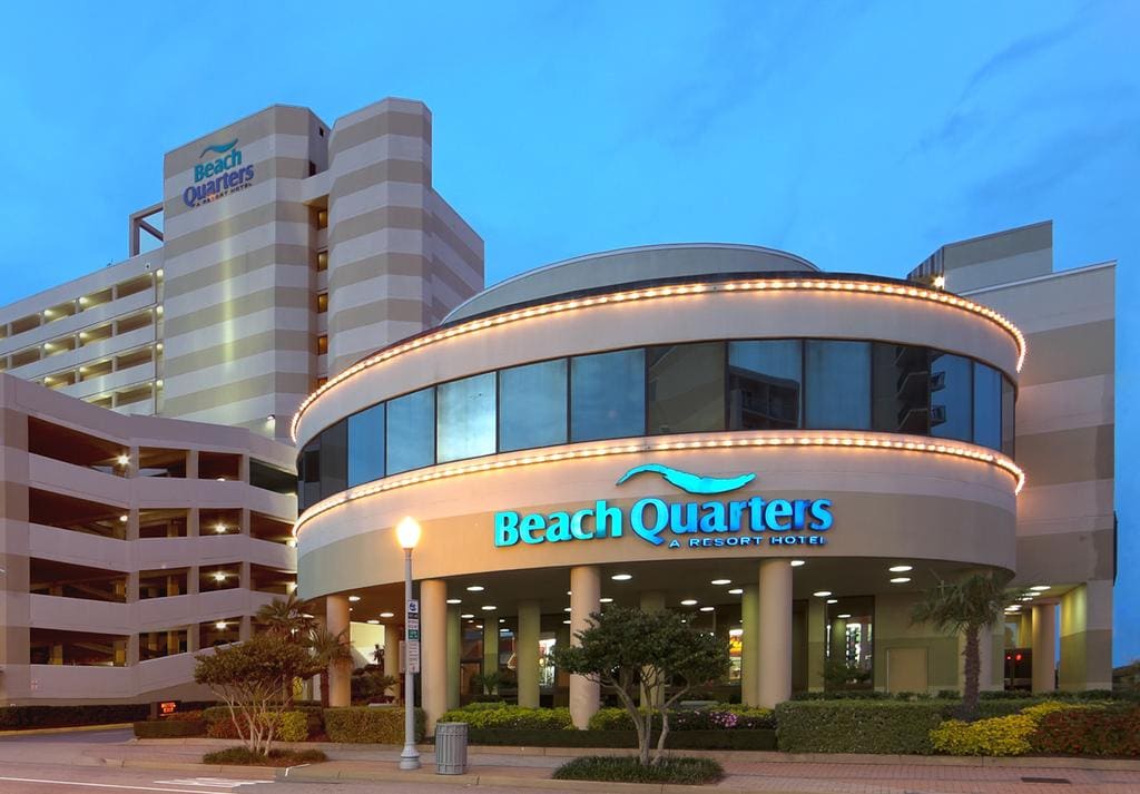 The exterior of Beach Quarters Resort is illuminated in the evening, showcasing its modern architecture. The entrance features curved windows and welcoming signage, while a surrounding area includes palm trees and landscaped greenery, creating an inviting first impression for guests.