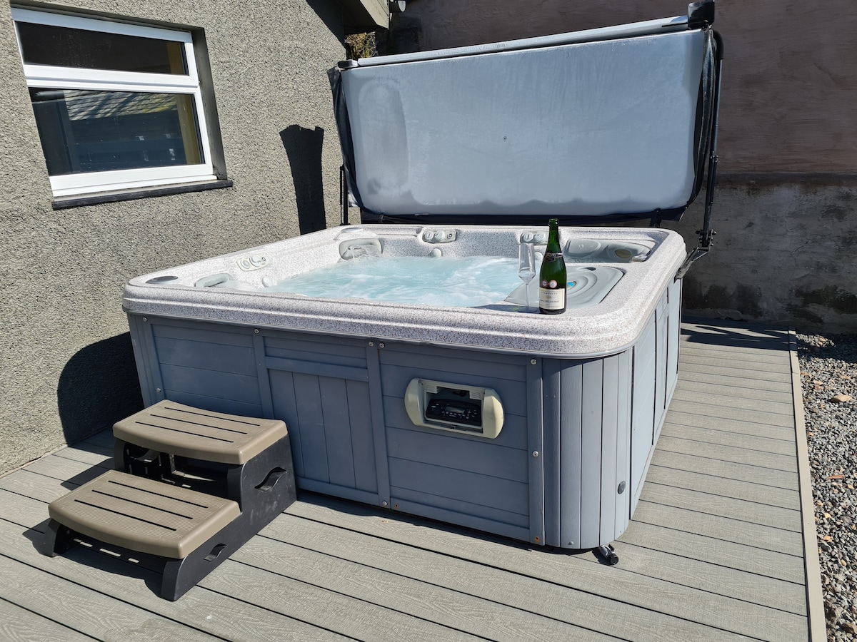 A five-person hot tub is situated on a wooden decking area, with a protective cover propped open. Two steps provide access to the tub, which features built-in jets and a small table holding a bottle, suggesting relaxation options are available during sunny moments.
