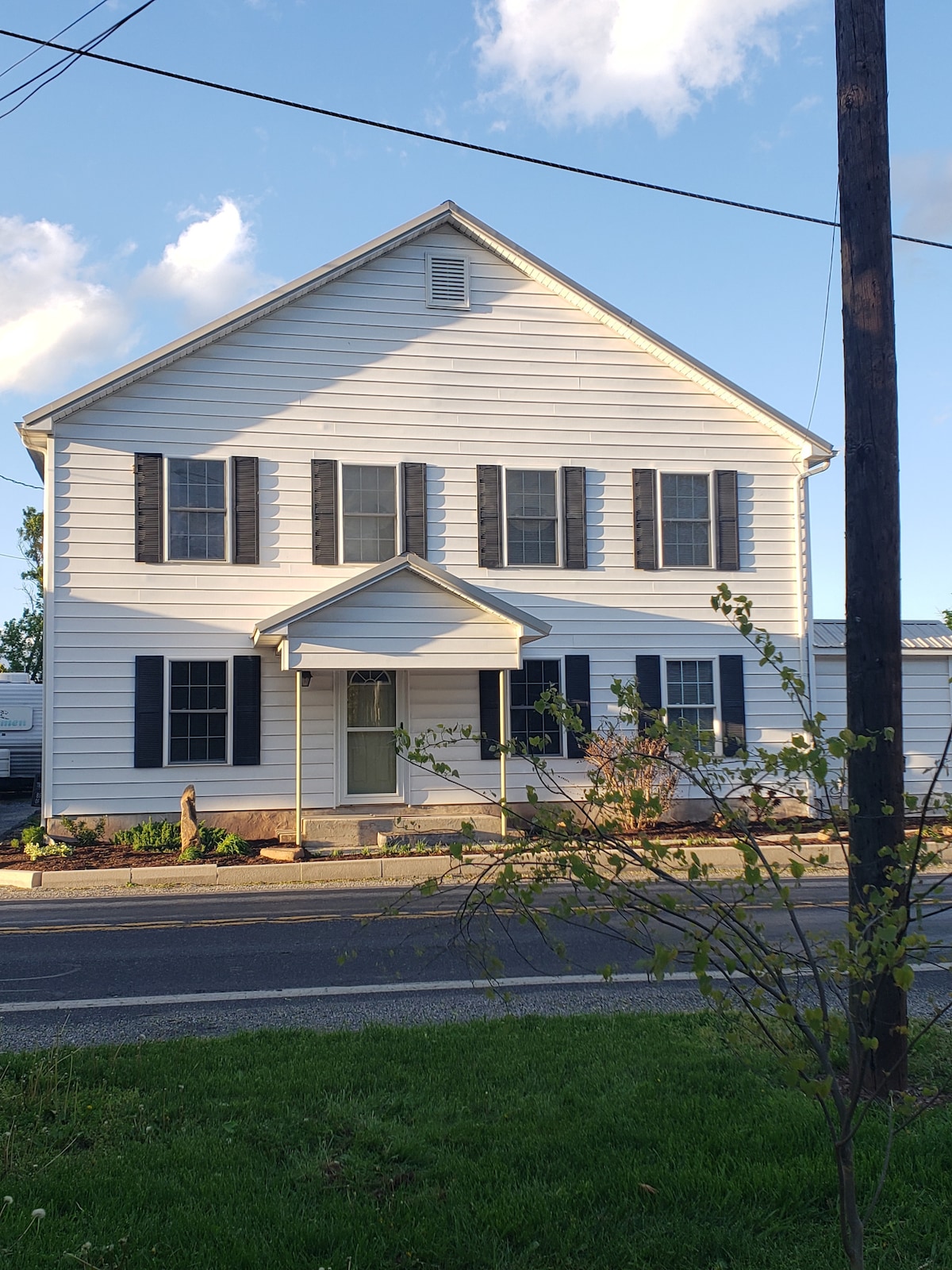 The exterior of a two-story house is presented, featuring white siding and dark shutters. A small front porch with a triangular roof is visible, along with neatly landscaped flower beds and a grass area in front. Power lines are seen in the background.