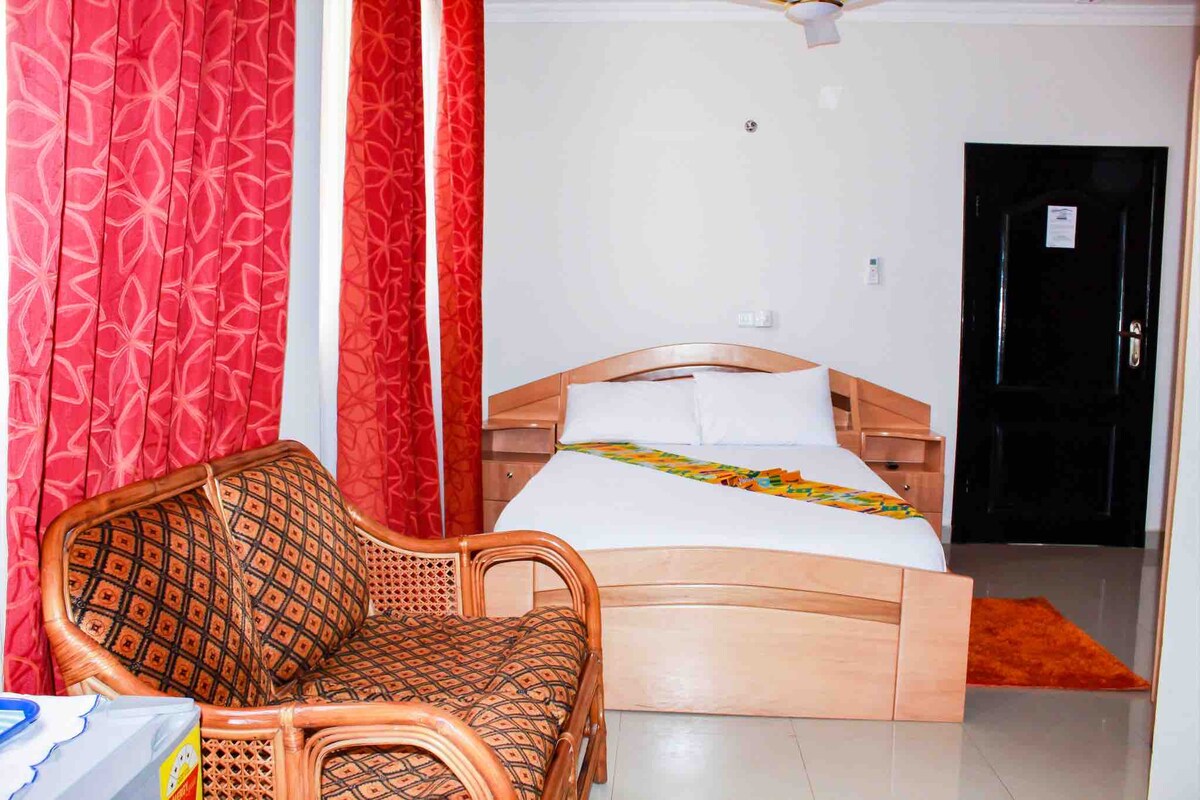 A well-lit room features a spacious bed with a colorful decorative element. A cozy seating area is provided by a woven chair set against a wall with rich red curtains. Light-colored walls create a bright atmosphere, accented by a dark entrance door.