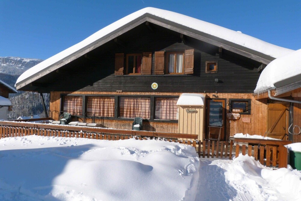 The chalet features a dark wooden facade with multiple windows framed in wood, partially covered with snow. The entrance is marked by a small shed, and outdoor seating can be seen arranged on the snow-covered porch, set against the backdrop of a clear blue sky.