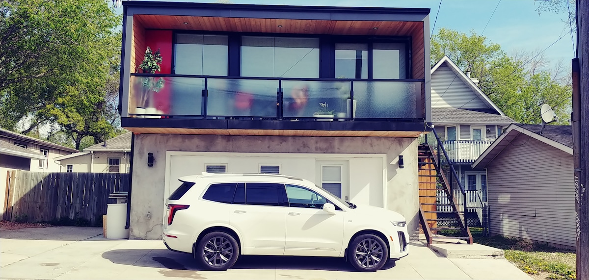 A modern guesthouse is showcased, elevated above a concrete garage. The upper level features large windows with a balcony, providing a view of the neighborhood. A white vehicle is parked in front, highlighting accessible parking just outside the entrance.