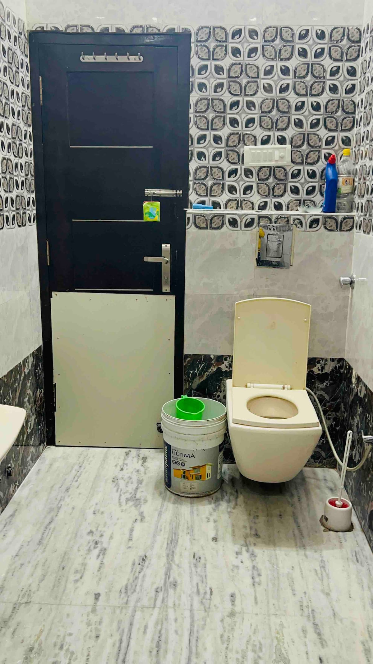The bathroom features a modern design with patterned tiles and a clean, bright appearance. A toilet is positioned beside a small bucket for waste disposal. The door is dark, contrasting with the lighter wall tiles, while a shower fixture is visible at one side.
