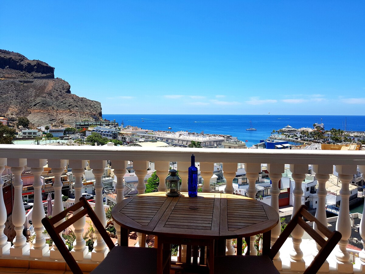 A private terrace is shown with a wooden table and two chairs positioned to overlook a stunning coastal view. The horizon displays a bright blue ocean under a clear sky, with distant boats visible and a rocky landscape framing the scenery.