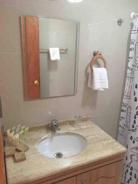 A bathroom featuring a light-colored stone countertop with a sink and a mirror above. Towel racks hold neatly arranged white towels. A shower curtain with a minimalist design adds a touch of style, while toiletries are presented in a wooden tray beside the sink.