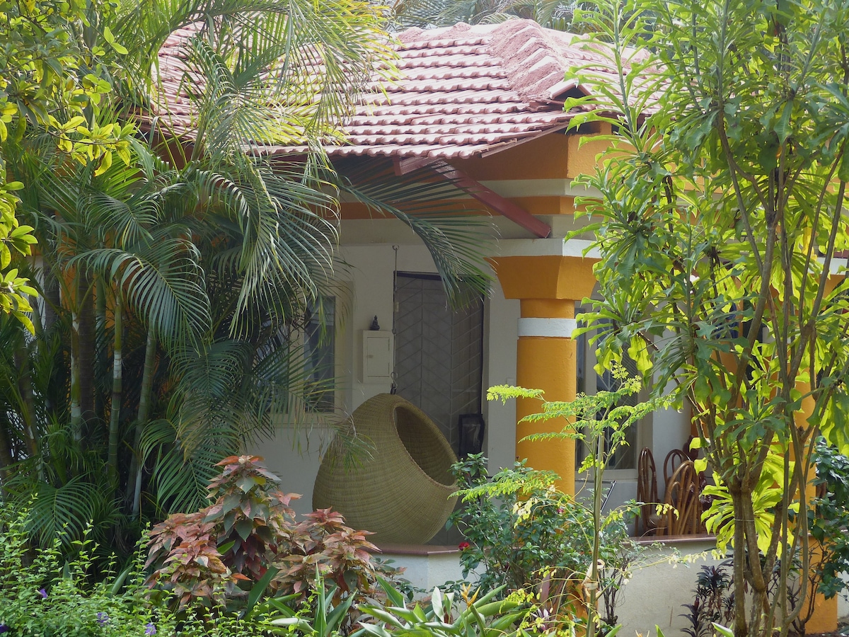 A charming entrance is framed by lush greenery, showcasing a mix of tropical plants and vibrant flowers. A unique woven chair is positioned under the porch, inviting relaxation. The structure features colorful pillars and a tiled roof, contributing to the cottage-like ambiance.