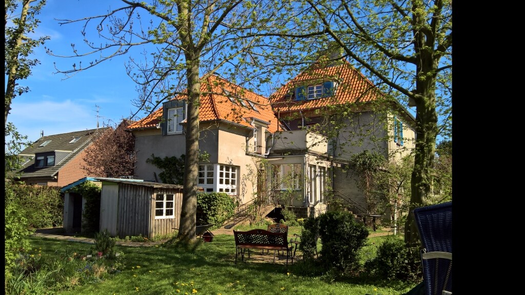 A house with a charming facade and a sloped orange-tiled roof, surrounded by greenery and trees. A wooden garden shed can be seen on the left, and a bench sits in the foreground, inviting relaxation in the spacious garden.