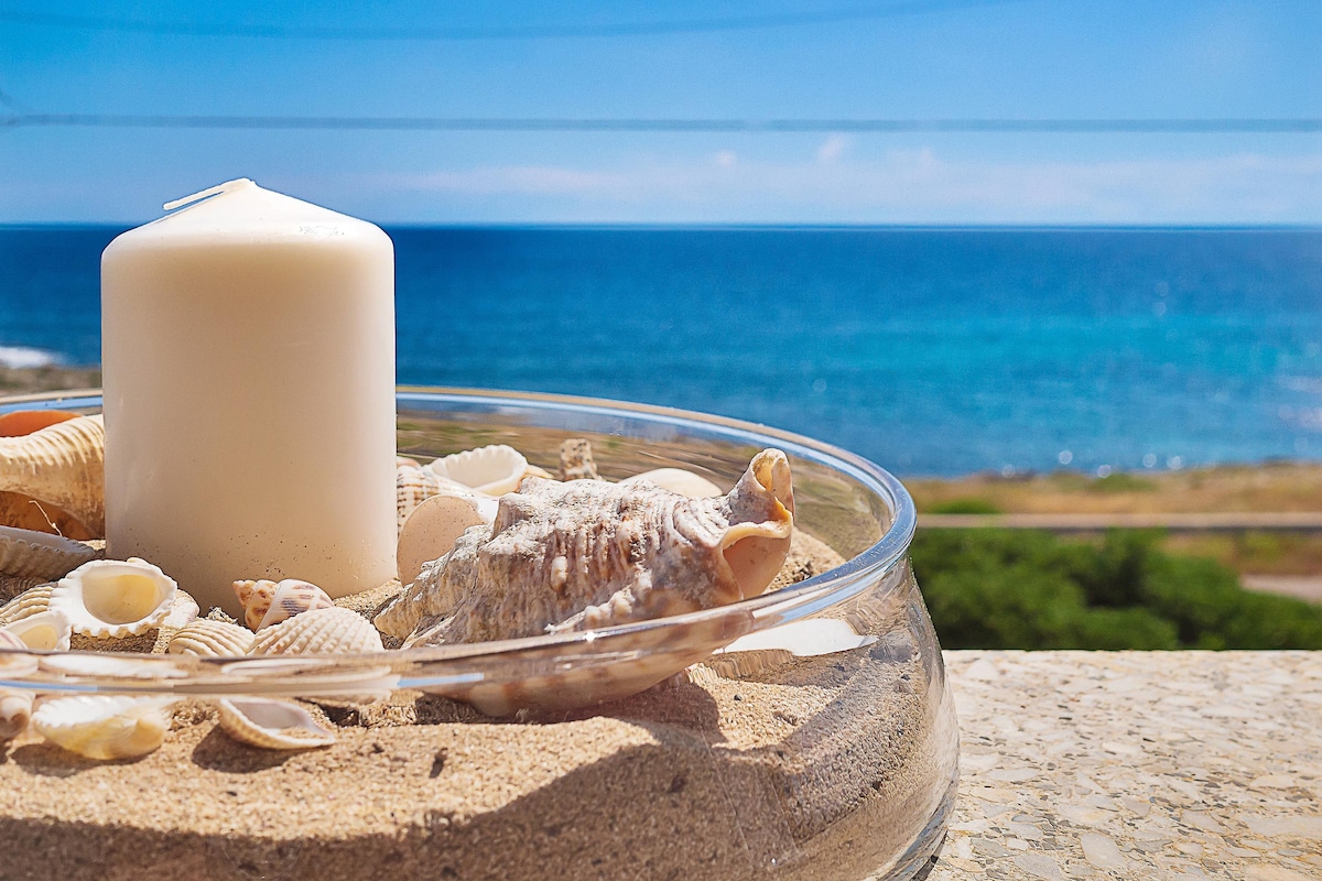A decorative bowl filled with sand and seashells is positioned on a surface. A white candle stands in the center, with the blue sea and clear sky visible in the background, creating a serene coastal ambiance.