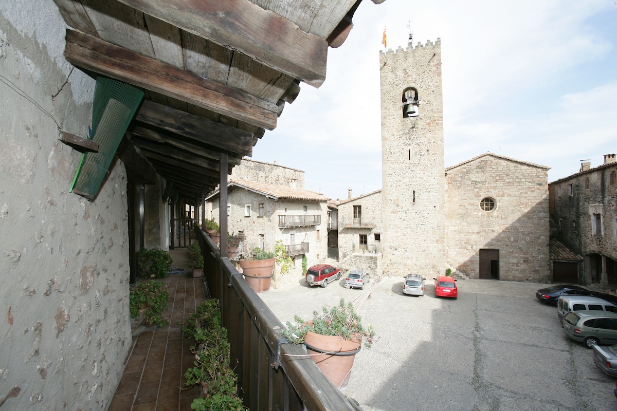 A view from a covered balcony showcases a charming courtyard with a stone tower, surrounded by rustic buildings. Potted plants add greenery to the space, while parked cars are visible on the gravel surface below. The sky is bright, enhancing the historical atmosphere.