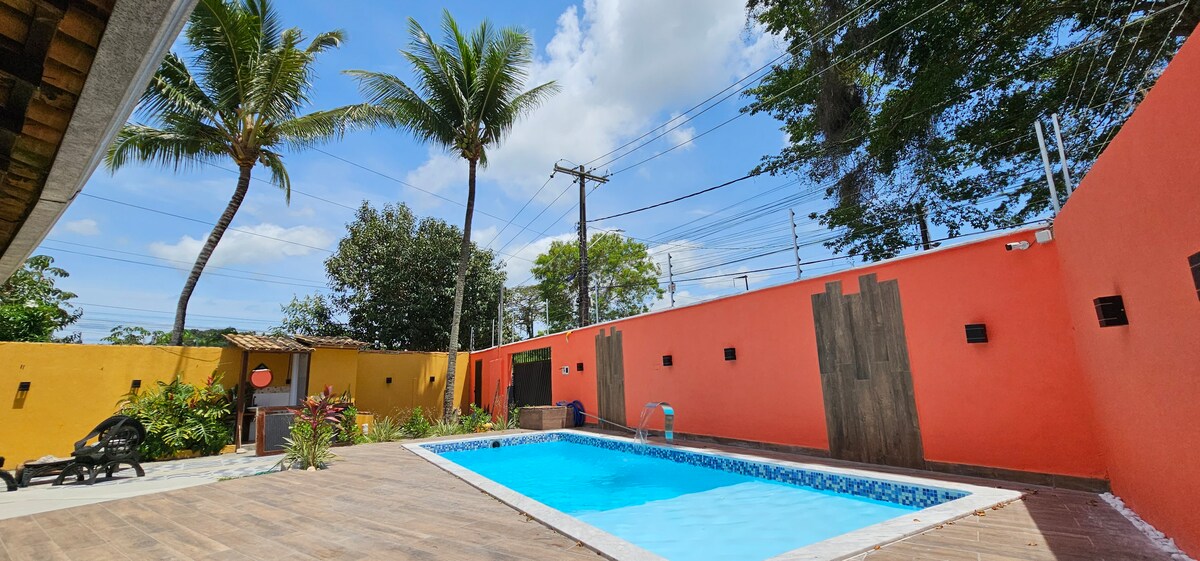 A clear blue swimming pool is surrounded by a wooden deck and vibrant walls in shades of orange and yellow. Lush palm trees provide shade against a sunny sky, while seating areas are visible nearby. The space emphasizes a tranquil outdoor atmosphere.