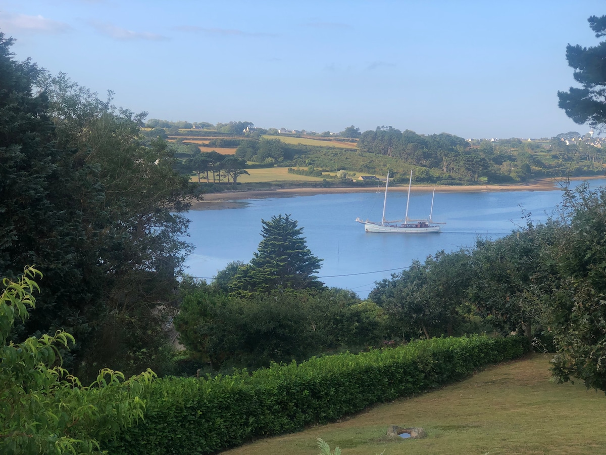 A serene view of the Aber is captured, featuring a sailboat gently resting on the water. Lush greenery and trees line the foreground, while rolling hills and fields create a picturesque background under a clear blue sky.